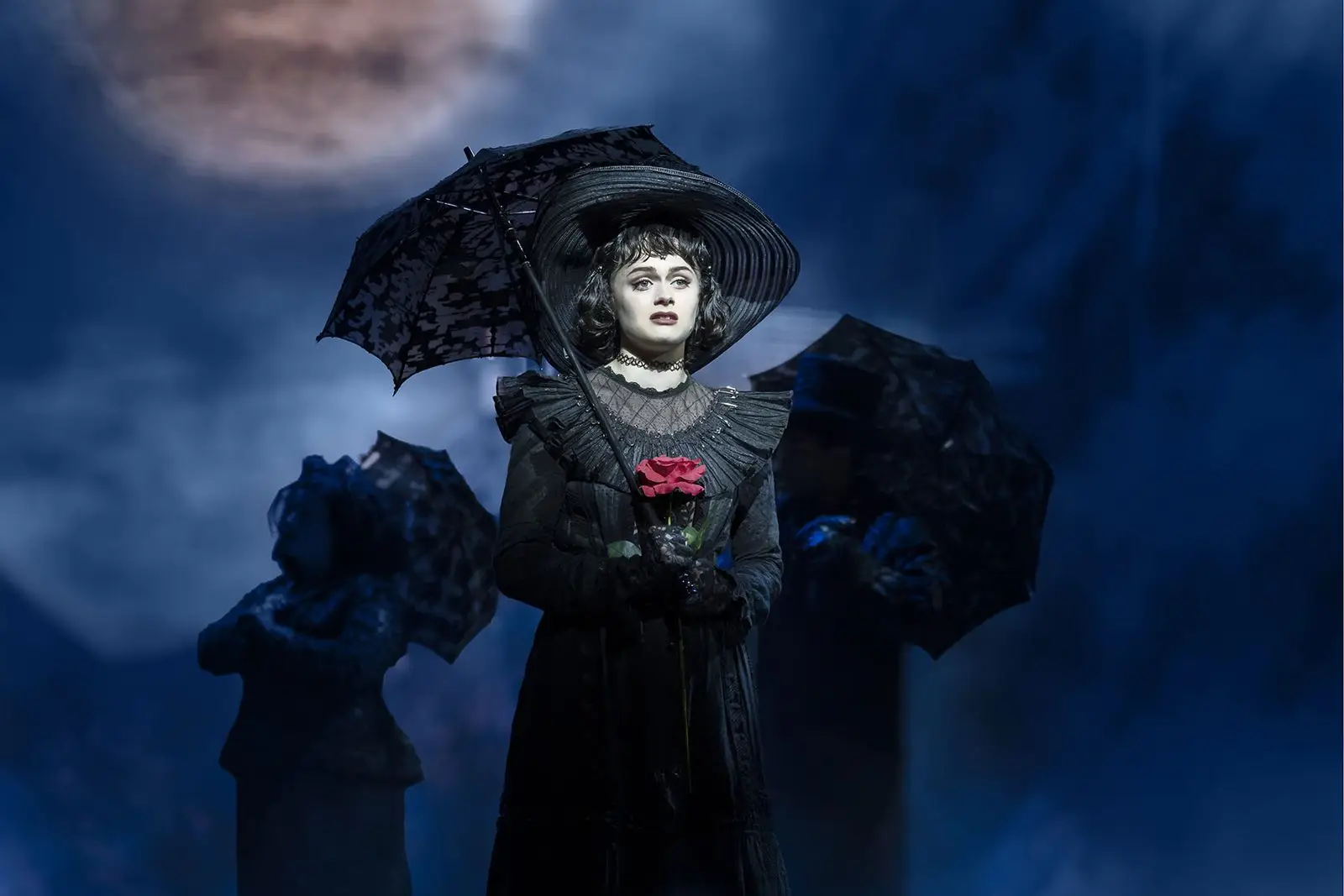 A black-clad figure with a parasol and red rose beneath a full moon in Beetlejuice The Musical at the Prince Edward Theatre - photography by Johan Persson